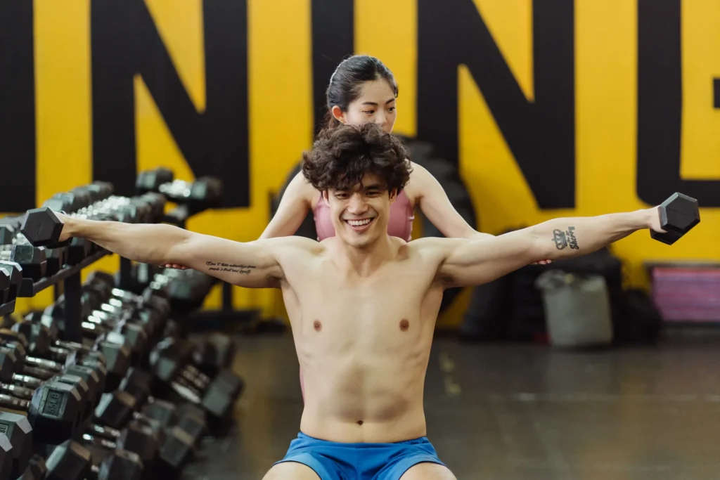 A man performs seated dumbbell lateral raises as part of side delt exercises while a trainer corrects his shoulder form